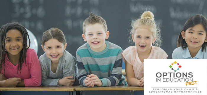 A multi-ethnic group of elementary age school children lean in on a row with their elbows on a desk as they smile and look at the camera.